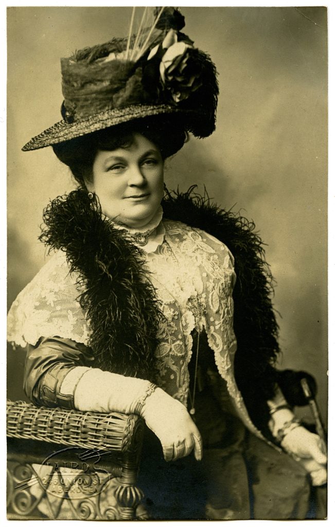 An archive photo of Jeannie Henry Bodie ‘Princess Rubie’ posing on a chair wearing a large hat and a feather boa.