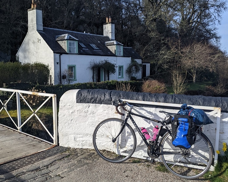 A touring bike laden with bags, against a wall on a canal bridge, with a historic loch keepers cottage by the bridge