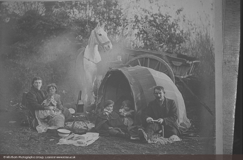 A gypsy family at an evening encampment: the father, mother and four children sit round a fire with cooking pots, while behind them is their horse and cart.