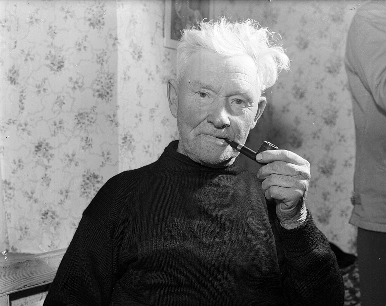 A white-haired man in a black knitted jumper draws on his pipe