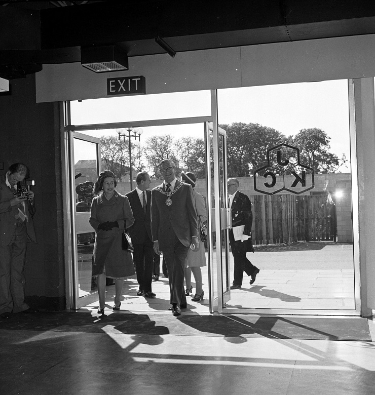 The Queen and Prince Philip, along withy various dignitaries, step through the glass doors of a leisure centre