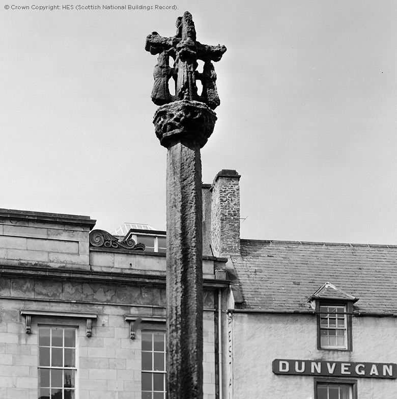 A carved medieval cross sits on a pillar about an historic town street.