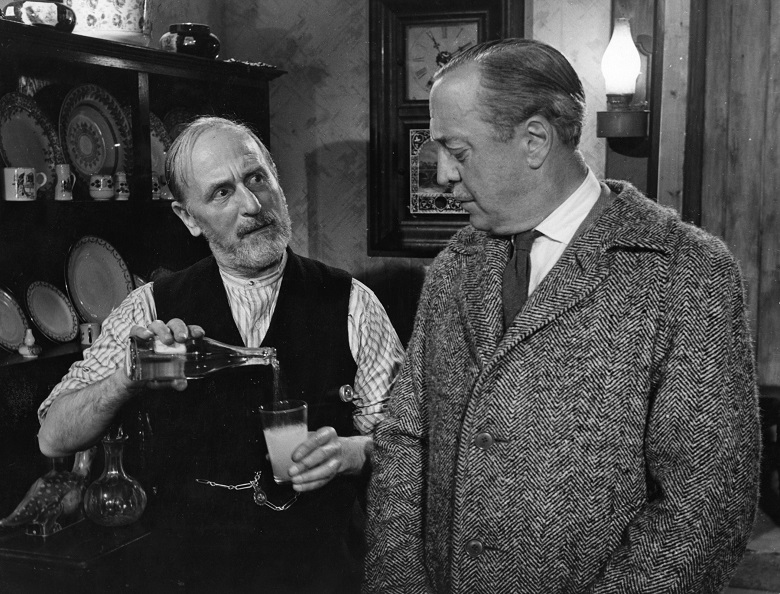 A still from a black and white film showing a man pouring a drink into a glass while another looks on