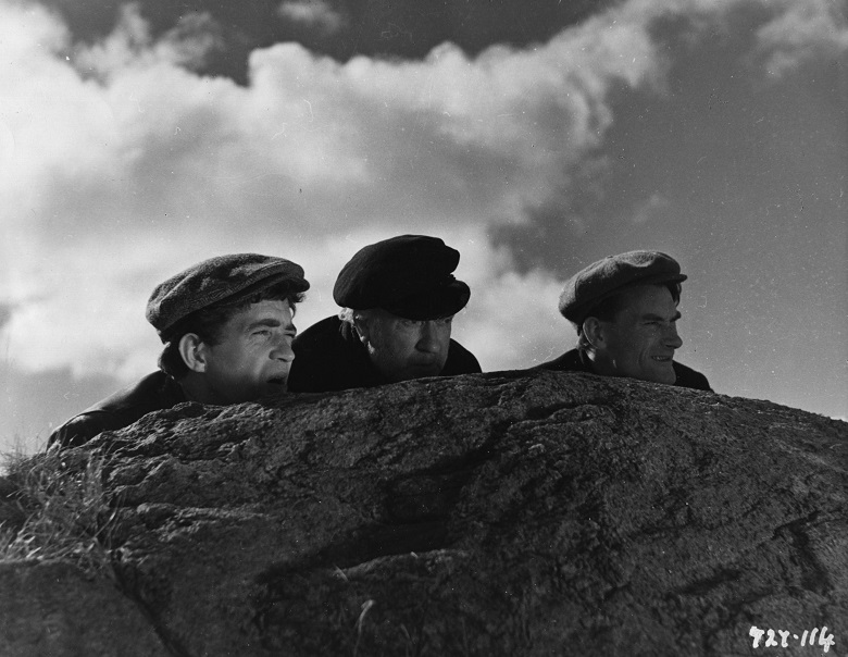 A still from a black and white film showing three men crouching behind a rock
