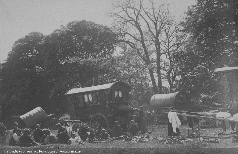 A group of men, women and children seated by uncoupled decorated gypsy caravans and covered wagons parked below trees.