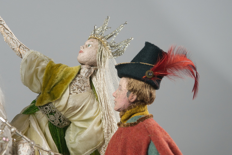 A close-up shot of two dolls. One depicts a fairy queen in a crown and gold dress, the other a man in a black hat adorned with a red feather