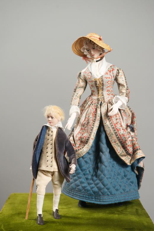 Small models of well dressed young boy with a walking stick alongside a lady in a colourful dress and sun hat