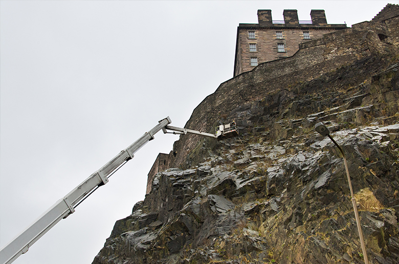 A long cherry picker carries workers up to castle rock to remove vegetation