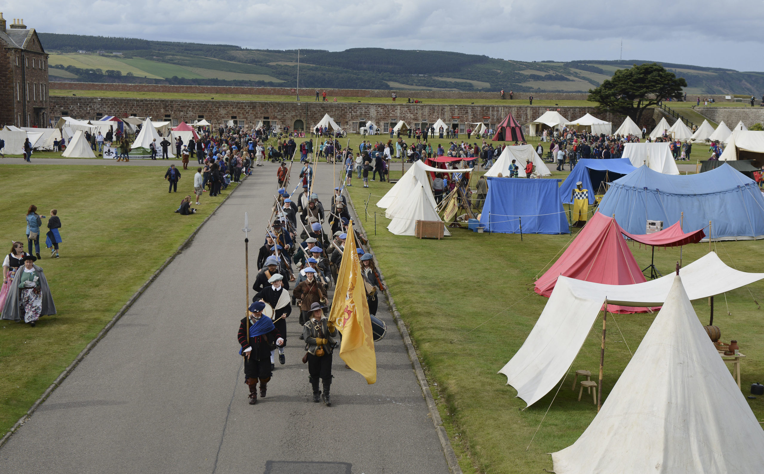A drone image of people dressed in re-enactment clothing carrying a flag and marching alongside festival tents for Celebration of the Centuries