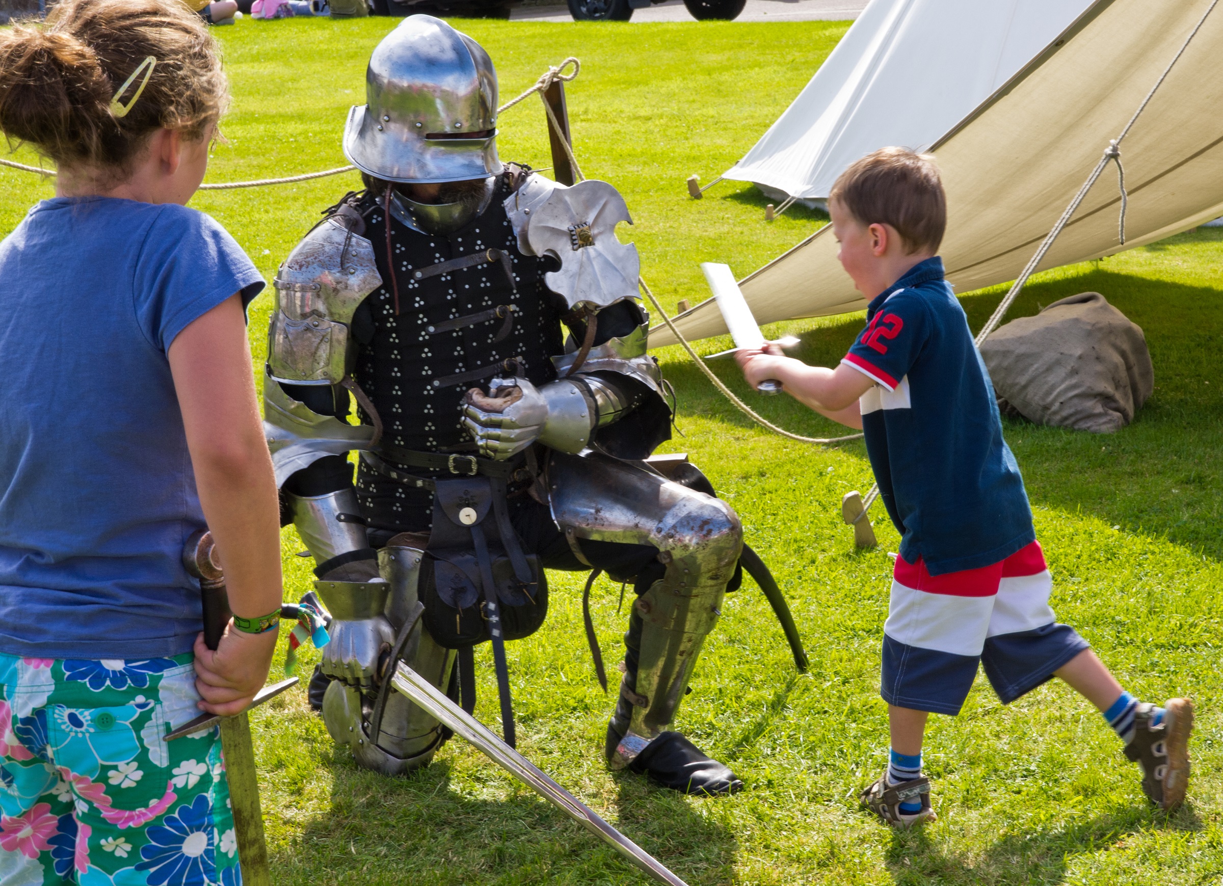 A person dressed in re-enactment clothes as a knight kneeling next to a child swinging a sword