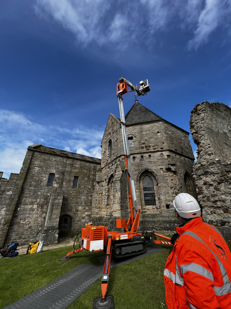 A person in a high vis jacket stands in front of an abbey with a cherry picker outside it