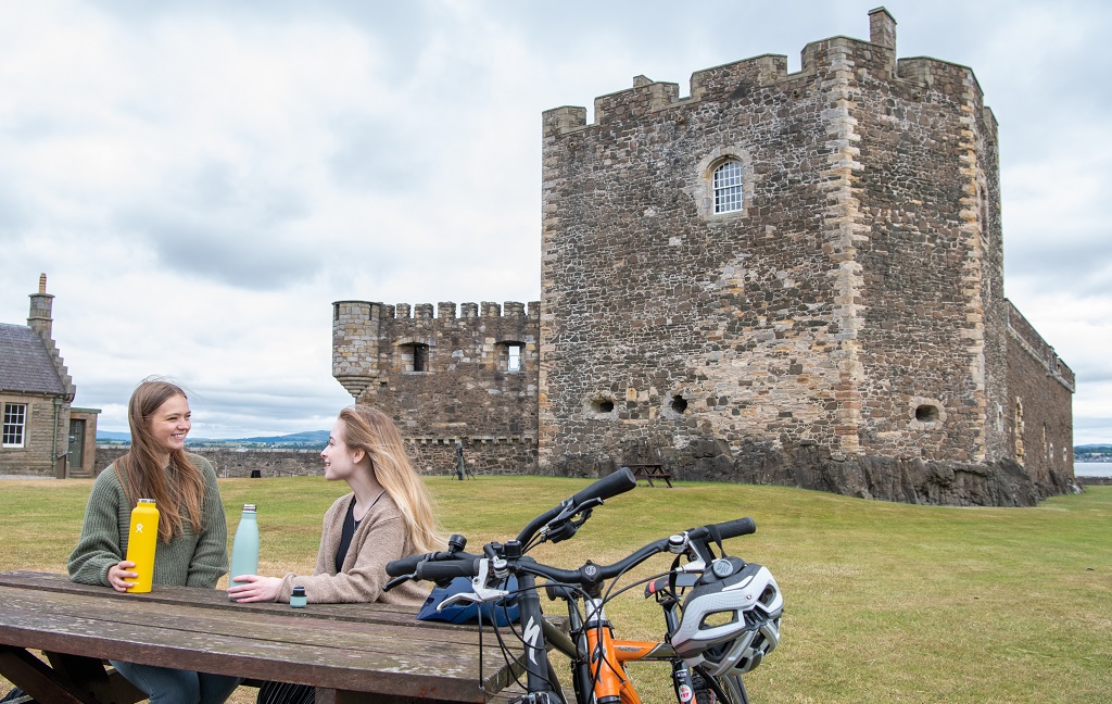 Two cyclists take a break at a picnic bench in front of a castle