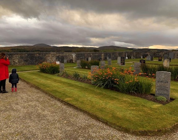 Standing in a cemetery, a woman in a red jacket is taking a photo with a toddler girl standing beside her.