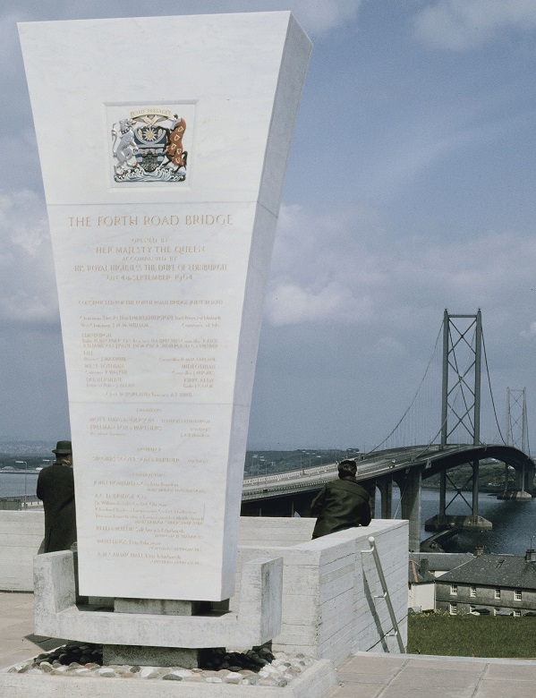 A commemorative monument made from smooth stone inscribed with with gold letters and royal insignia, explaining that the bridge in the background was opened by the Queen 