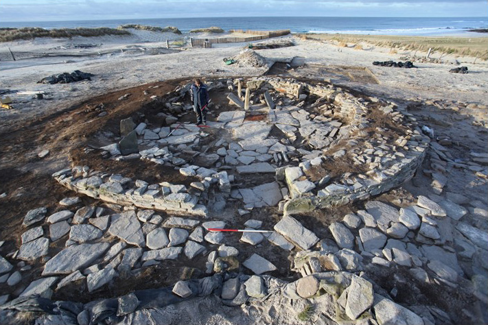 Remains of a circular, stone house. The stone is almost white. Inside the house a person stands in dark overalls. The house has no roof and the walls come to knee height and have brown soil on top.