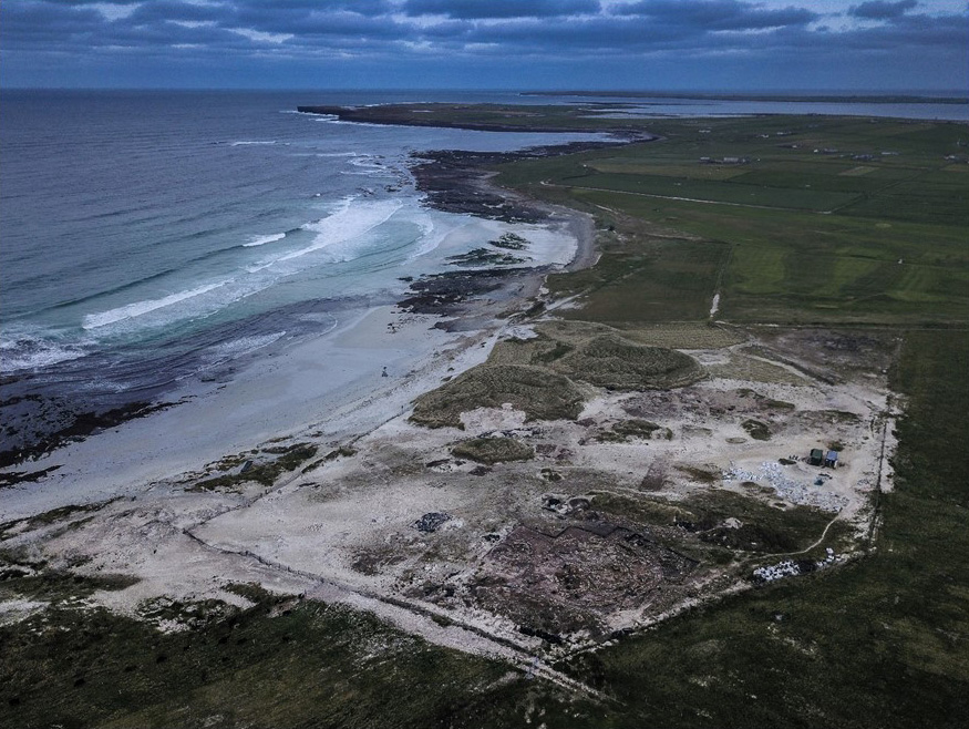 Aerial view of the dig site at the Links of Noltland in the sand, with beach on the left. The waves are small and the time of day is dusk