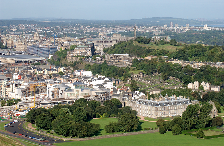 A view of the city of Edinburgh with a historic palace and the modern parliament building located close together in the foreground 