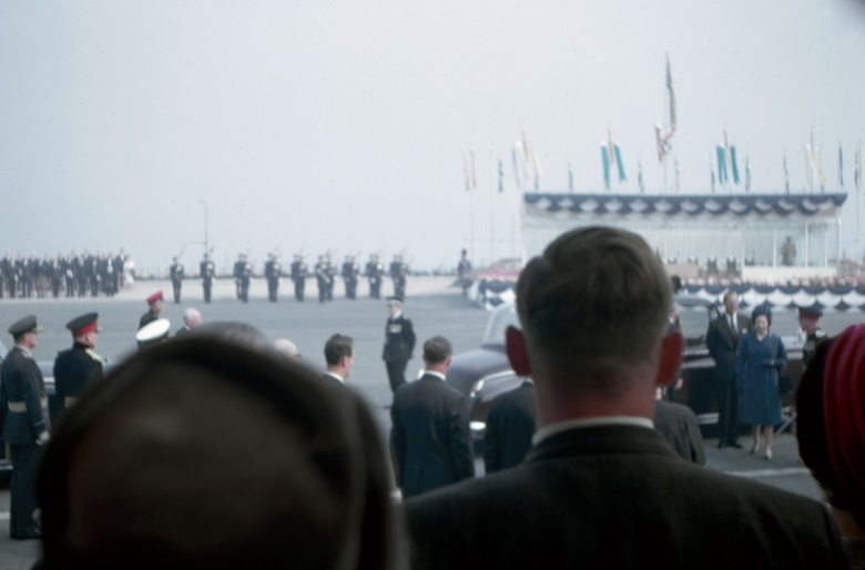 A view of the Queen arriving at an opening ceremony with various soldiers and dignitaries looking on 
