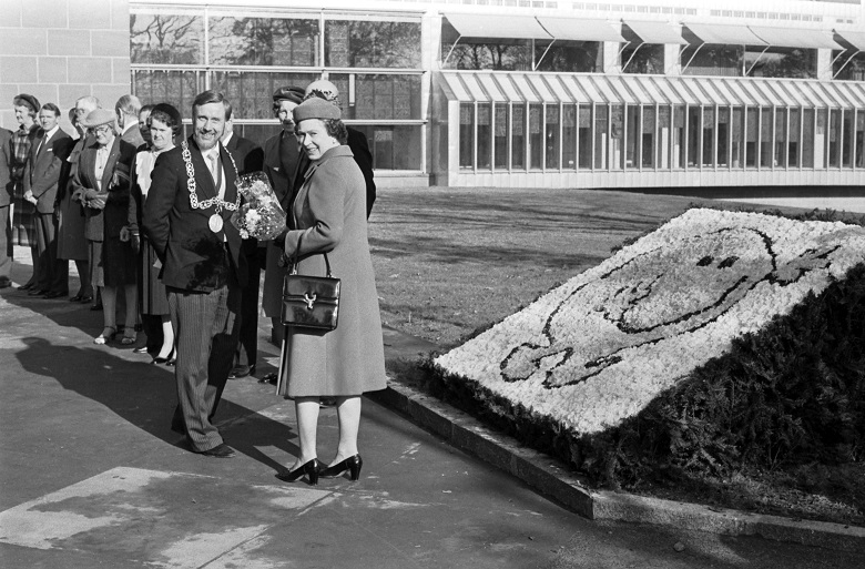 Archive photo of the Queen at the opening of a new museum. The Queen is standing beside a flower bed arranged into a smiley face motif.  