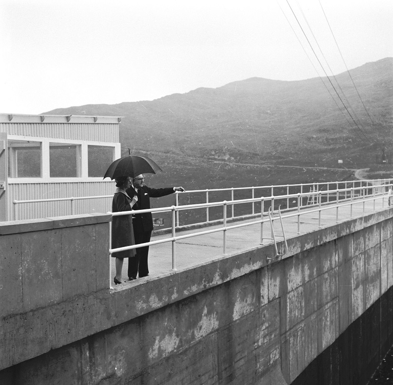 A black and white archive photo of the Queen with an offical on top of a large dam. She is sheltering under an umbrella. 