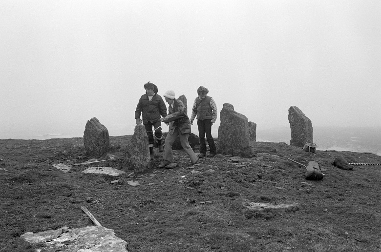 Three people in 1980s clothing walk across a chambered cairn.