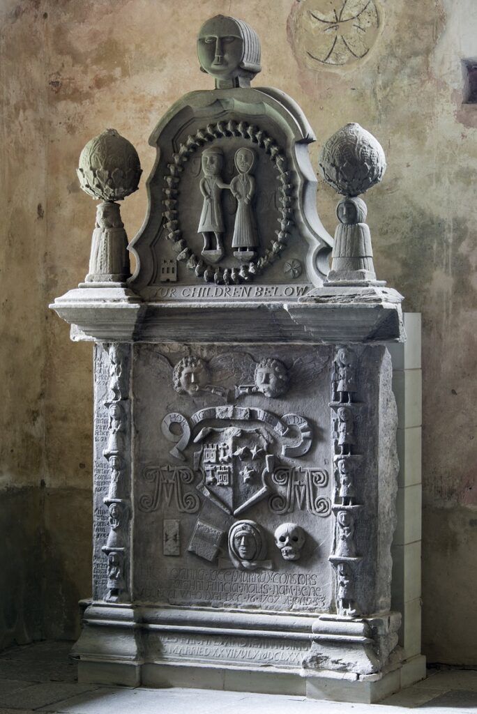 elabourately carved monument featuring a husband and wife carbed in the middle, their numerous children creating a border to the left and right of the stone and heraldic symbols.