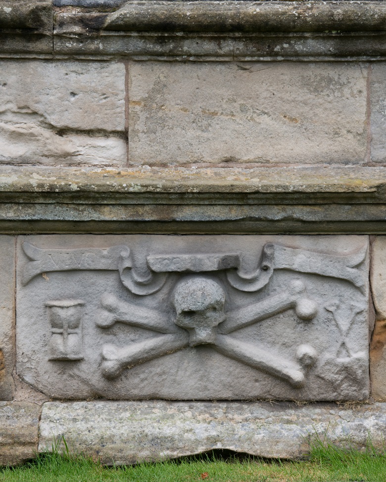 detail of a carved gravestone featuring a skull and crossed bones in the middle with an hour glass to the left