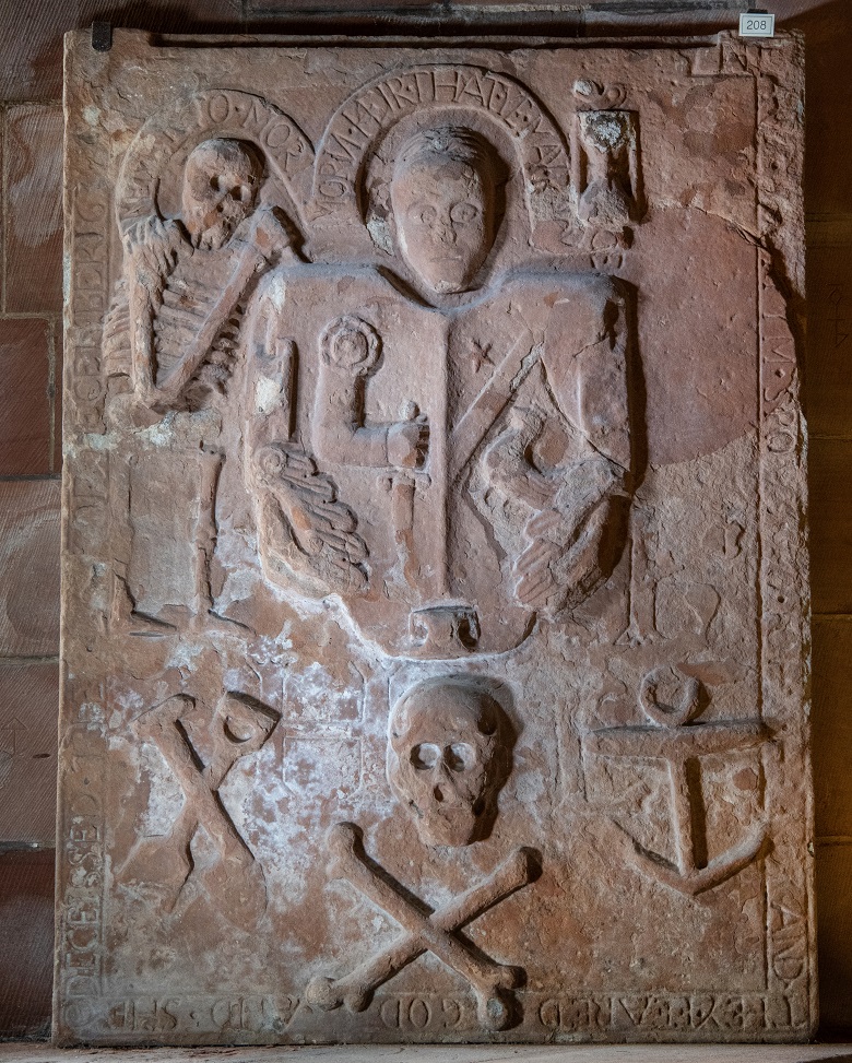 A carved gravestone featuring a heraldic panel in centre with memento mori symbols around the edge, including a skeleton, skull and cross bones and hour glass.