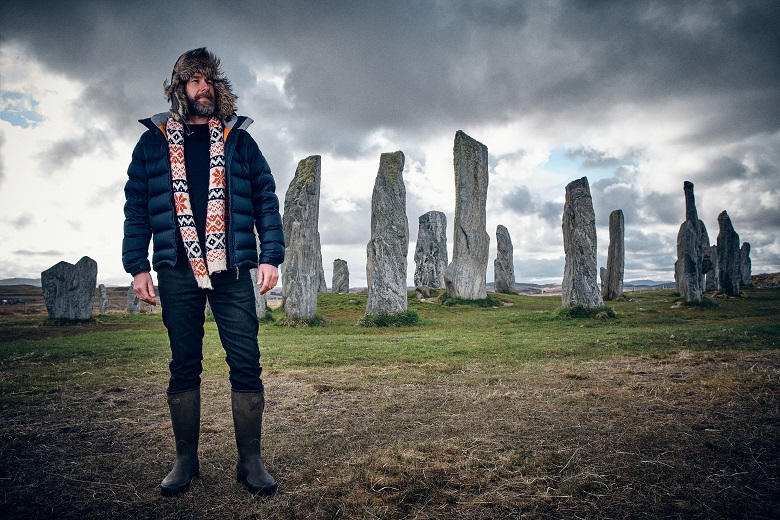 The Hebridean Baker, wearing a colourful knitted scarf and a fluffy hat, standing in front of a collection of standing stones