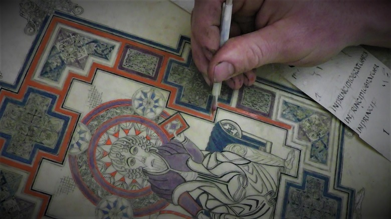 A close-up photo of an artist using a stylus to add colour to a reproduction of the Book of Kells