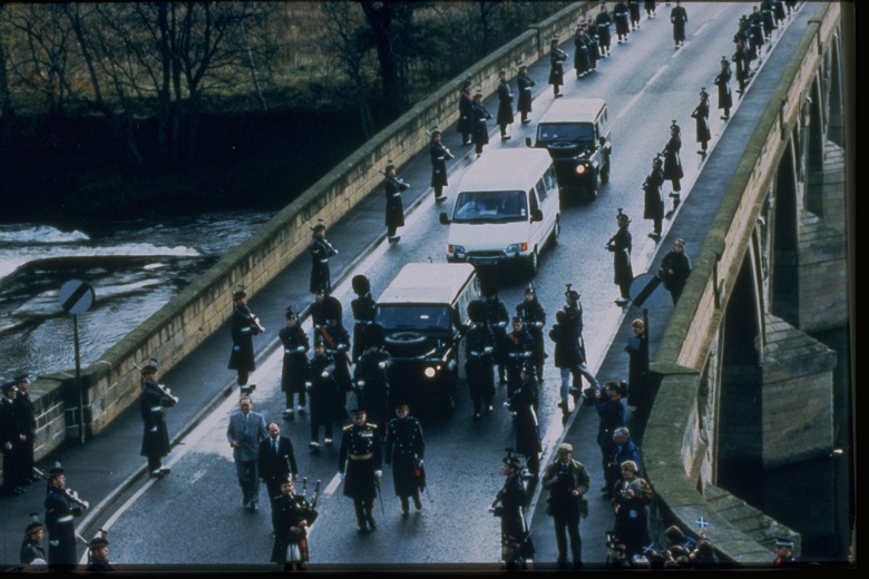 Pipers lining the road as a convoy of vehicles crosses a historic stone bridge