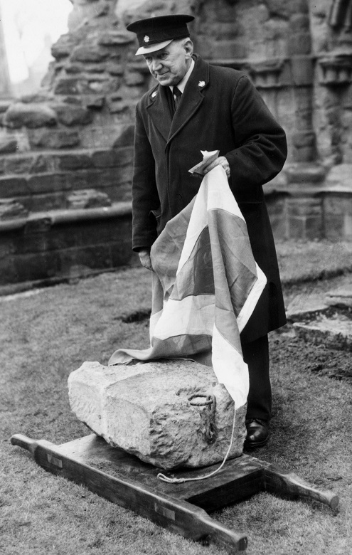 A black and white archive photo of a uniformed custodian standing over the Stone of Destiny within the ruins of an abbey. He holds a Saltire flag which appears to have been draped over the stone. 