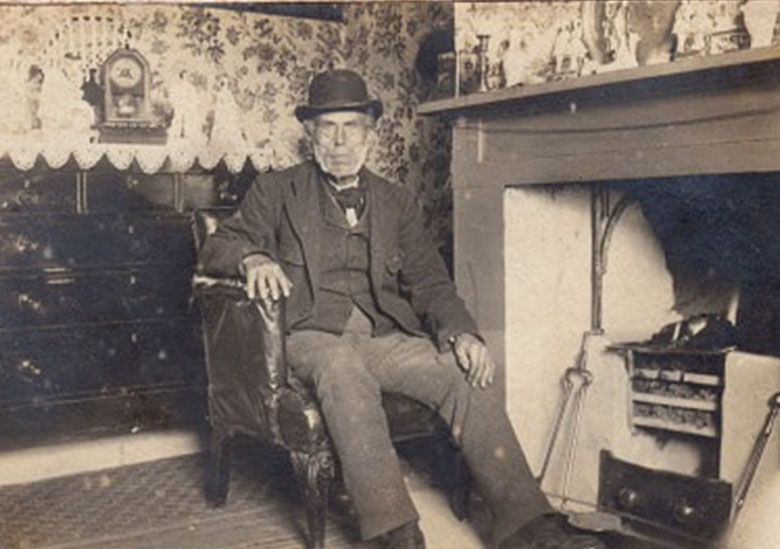 a man in a bowler hat sits in a cosy parlour in front of a fireplace