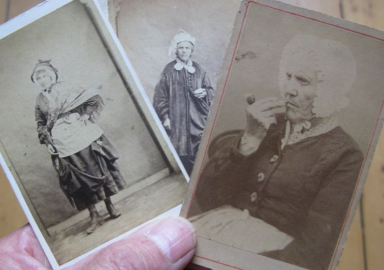a hand holding three photos of an older woman in Victorian clothing. In the top photo, she is smoking a pipe.