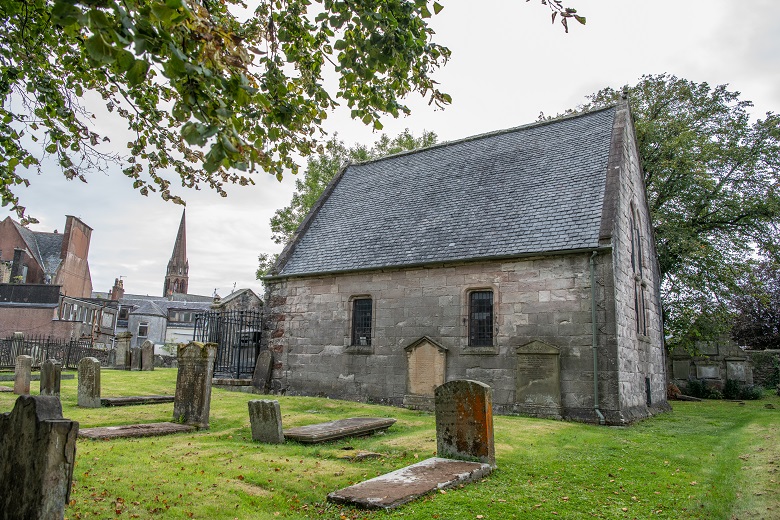 A small chapel with a plain exterior set in a graveyard. The skyline of Largs with a church steeple can be seen in the background.