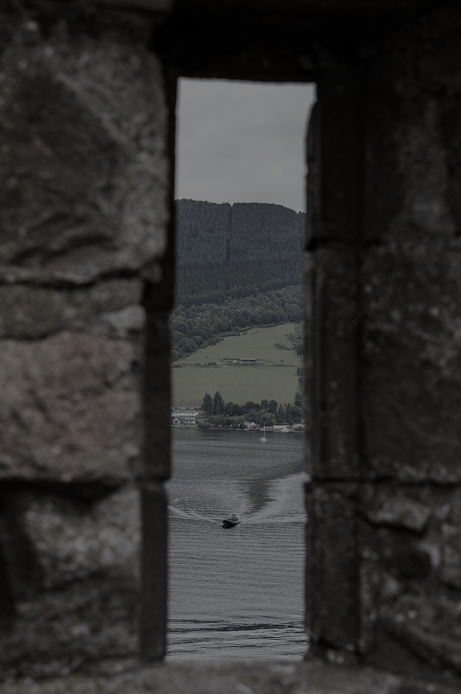 A view looking down on a boat a loch through a narrow, slit-like window at a castle