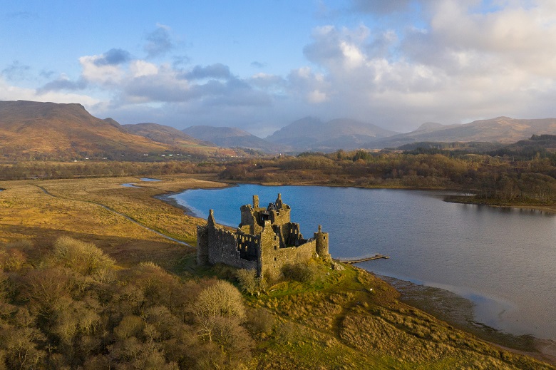 An aerial photo of a ruined castle beside a loch, surrounded by mountains