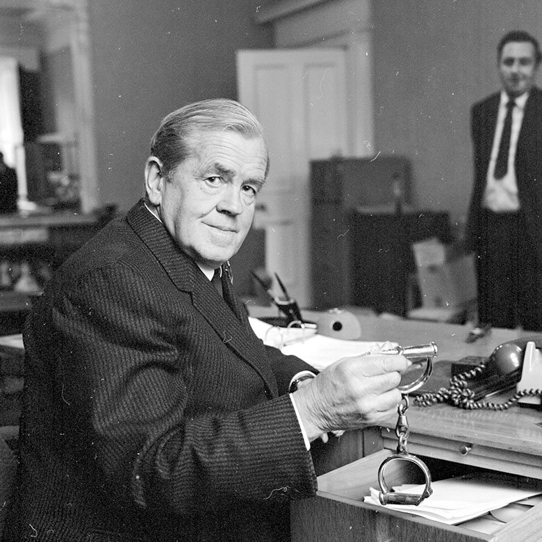 A man in a suit sits at an oldfashioned desk. He is lifting a pair of handcuff out of the top drawer.