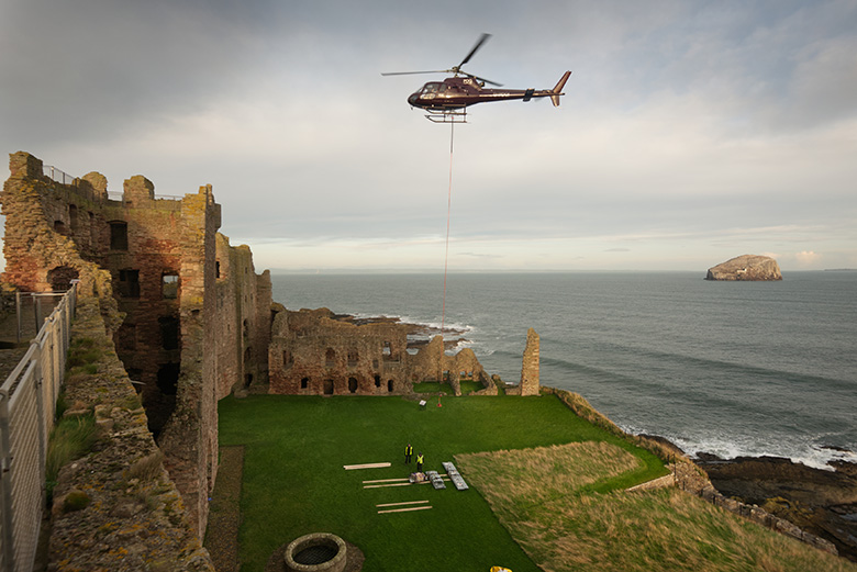 A helicopter hovers over the courtyard at Tantallon Castle delivering equipment on a rope. The sea and the Bass Rock are in the background.