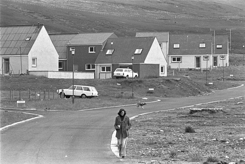 A black and white archive photo of a lady walking along a road away from newly built 1970s houses in a village in Shetland