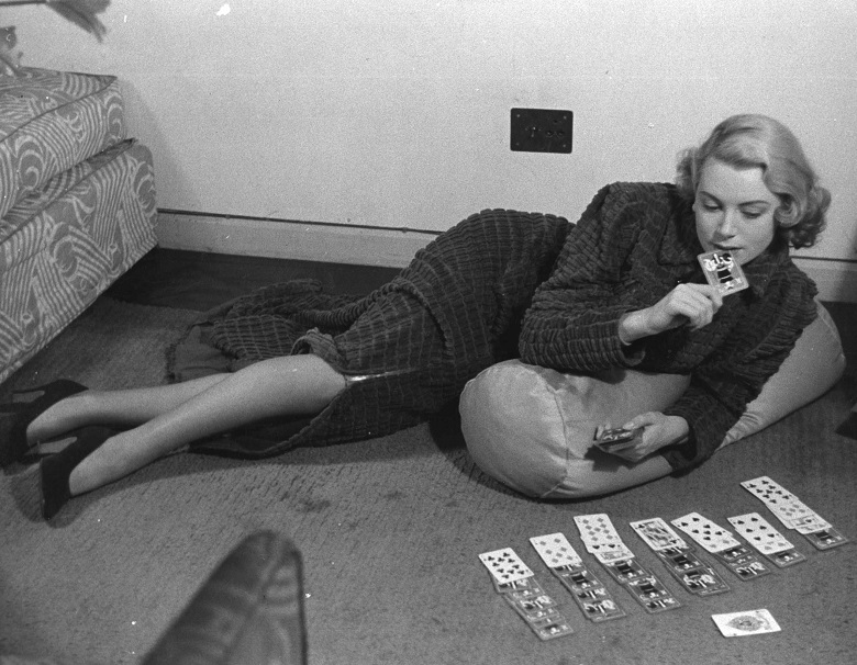 A lady in a reclining position holds a playing card to her lips while studying an assortment of cards laid out on the carpet in front of her