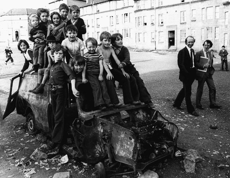 A black and white archive photo of two men in suits walking past a group of children climbing on a burnt out van