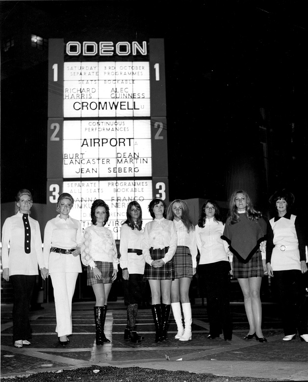 A group of ladies pose in front of a new Odean cinema with a brightly lit sign advertising upcoming films