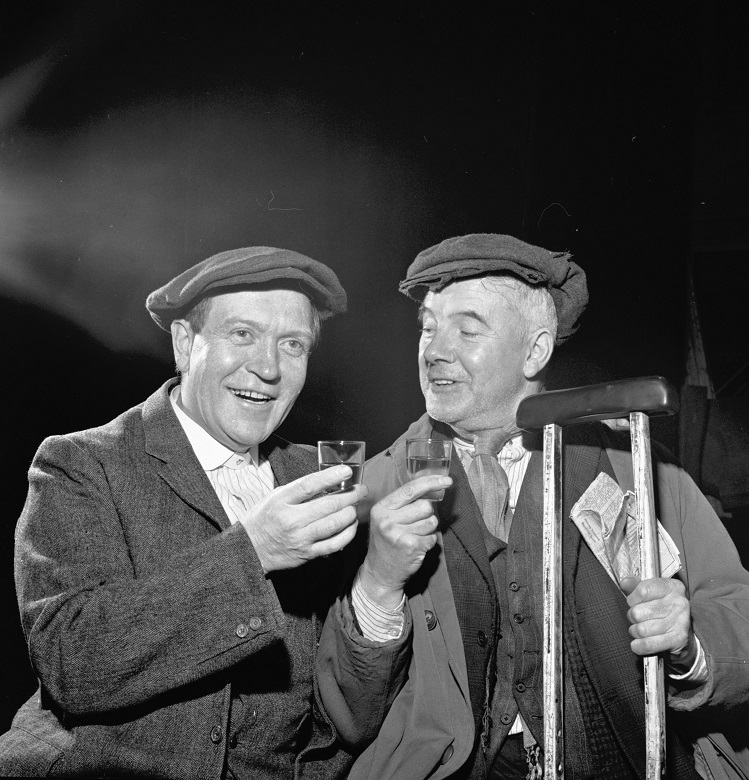Tow actors in flat caps share a dram of whisky during a play