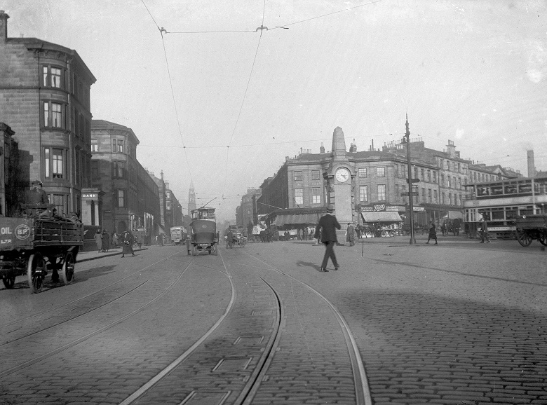 Black and white photo showing Edinburgh's Haymarket junction in the 1930s. The street is cobbled and there are tram lines. There are also carts and horses using the road. There is a war memorial in the junction and people in 1930s dress cross the road.