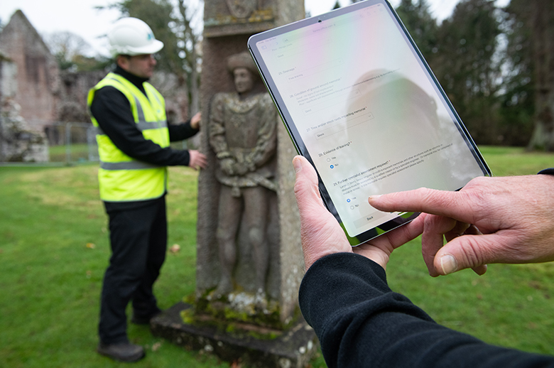 a person in high vis and a helmet inspects a carved gravestone. SOmeone in the foreground holds an iPad and makes notes.