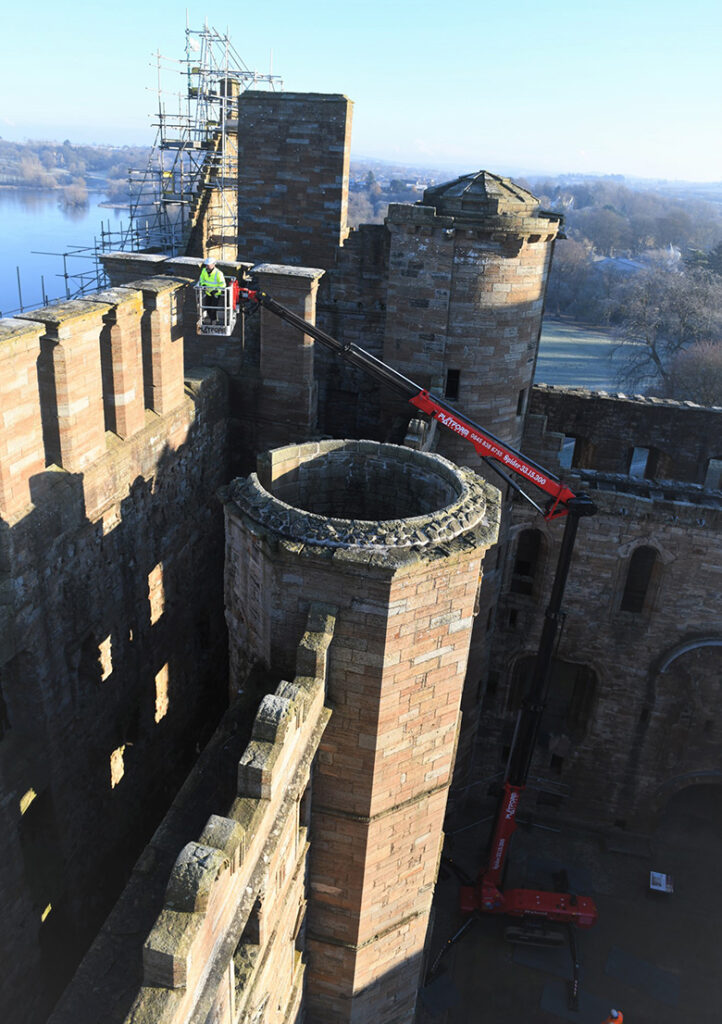 A person in the basket of an elevated platform checking the stonework at the top of Linlithgow Palace. The loch is in the background. It is a bright, frosty day.