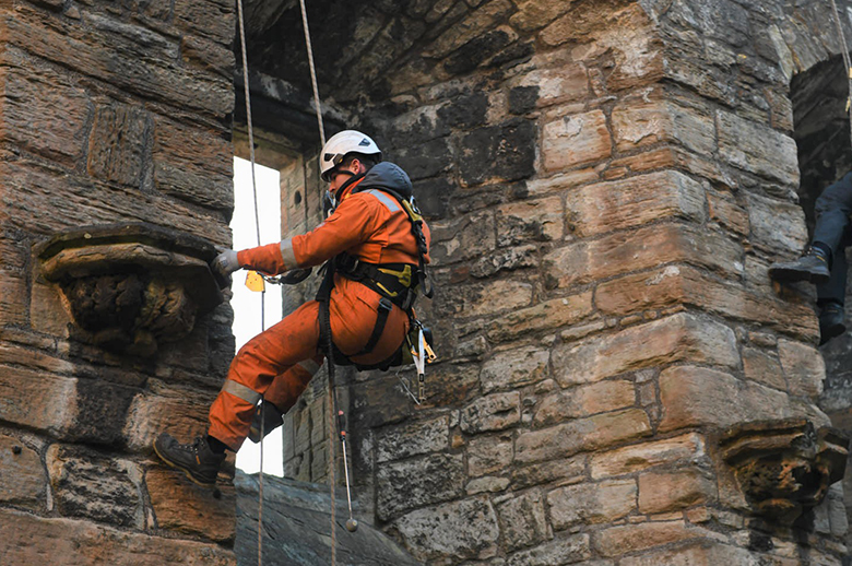 A person in an orange jumpsuit and hard hat hangs on an access rope in front of a stone wall. They are checking a piece of carved stonework with their hand.