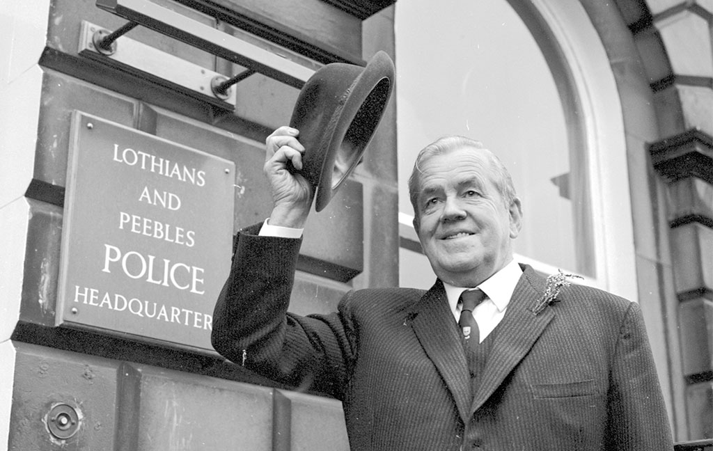 A man in a tweed suit raises his trilby hat. A sign behind him reads: Lothians and Peebles Police Headquarters