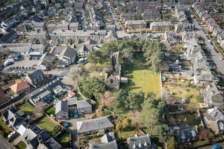 An aerial view of the ruins of a kirk, which is in a space nestled between gardens in a historic town.
